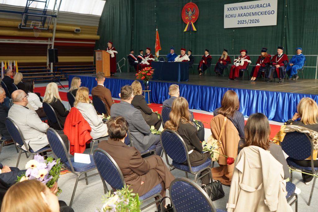 Osoby przybyłe na inaugurację siedzą na krzesłach ustawionych w rzędy na hali sportowej, w tle scena