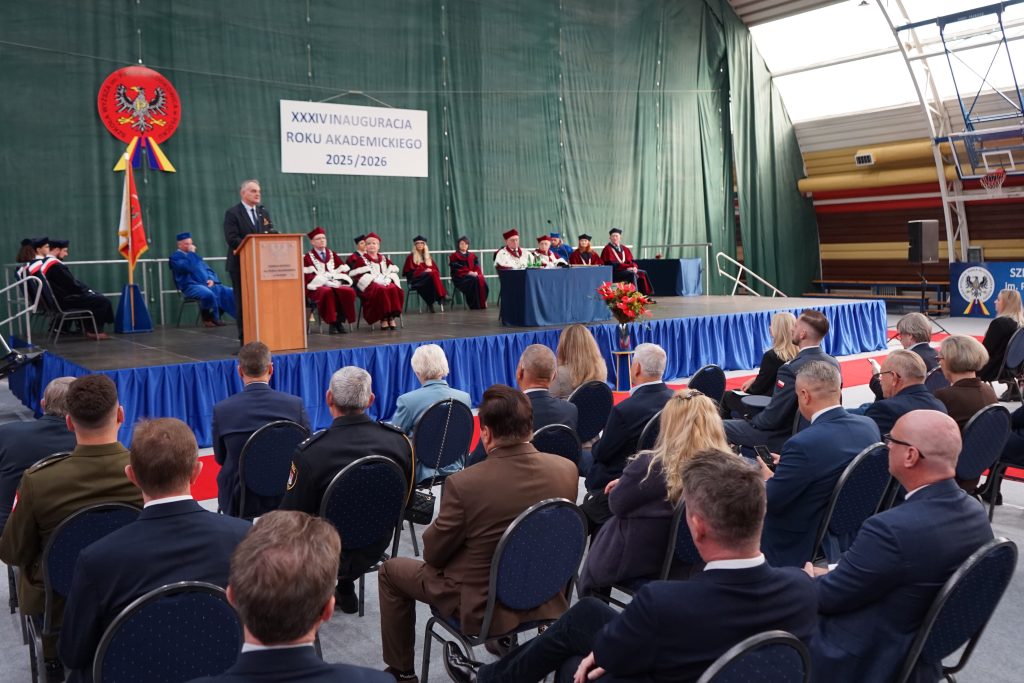 Osoby przybyłe na inaugurację siedzą na krzesłach ustawionych w rzędy na hali sportowej, w tle scena