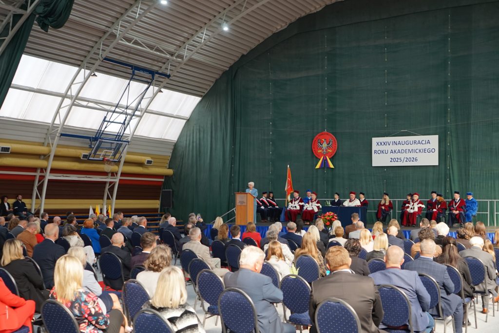 Osoby przybyłe na inaugurację siedzą na krzesłach ustawionych w rzędy na hali sportowej, w tle scena
