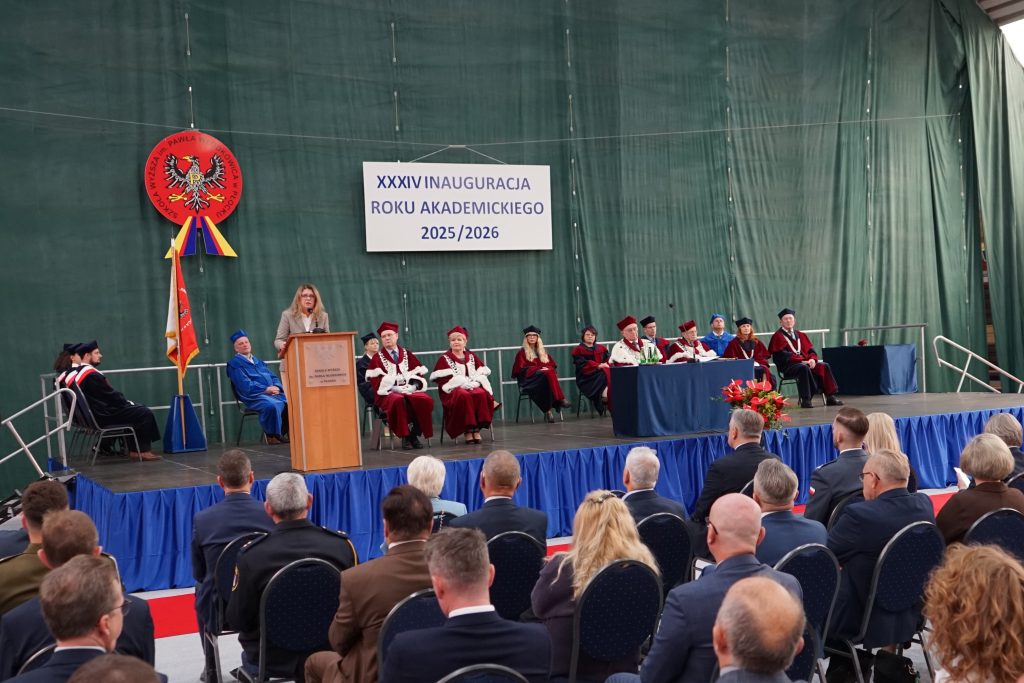 Osoby przybyłe na inaugurację siedzą na krzesłach ustawionych w rzędy na hali sportowej, w tle scena