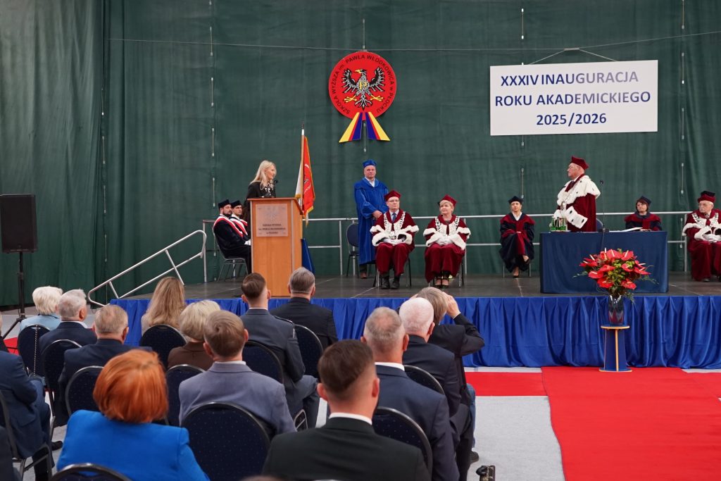Osoby przybyłe na inaugurację siedzą na krzesłach ustawionych w rzędy na hali sportowej, w tle scena