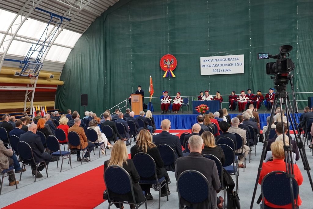 Osoby przybyłe na inaugurację siedzą na krzesłach ustawionych w rzędy na hali sportowej, w tle scena