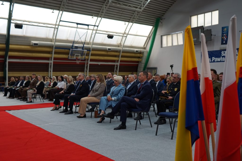 Osoby przybyłe na inaugurację siedzą na krzesłach ustawionych w rzędy na hali sportowej