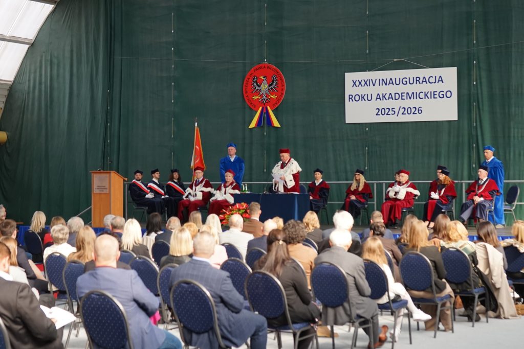 Osoby przybyłe na inaugurację siedzą na krzesłach ustawionych w rzędy na hali sportowej, w tle scena