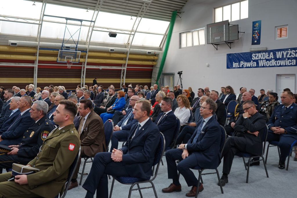 Osoby przybyłe na inaugurację siedzą na krzesłach ustawionych w rzędy na hali sportowej