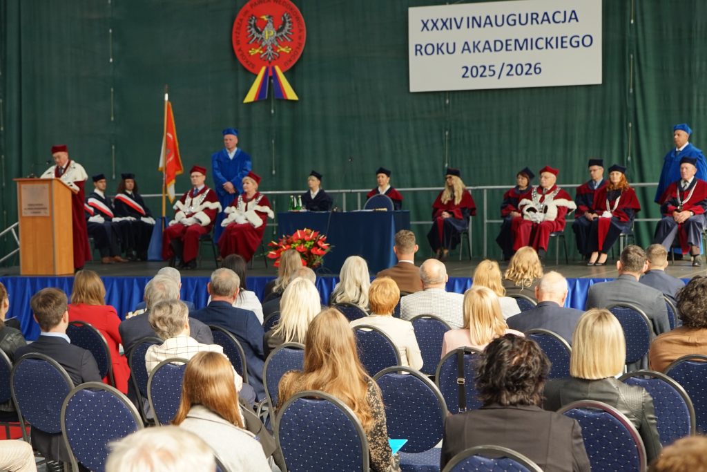 Osoby przybyłe na inaugurację siedzą na krzesłach ustawionych w rzędy na hali sportowej, w tle scena