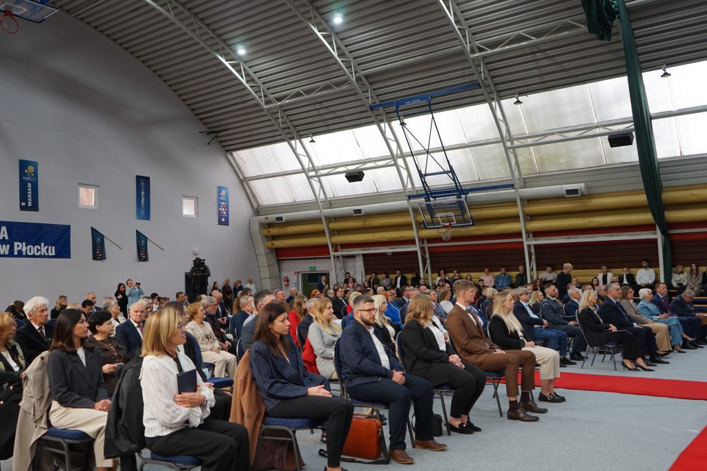 Osoby przybyłe na inaugurację siedzą na krzesłach ustawionych w rzędy na hali sportowej