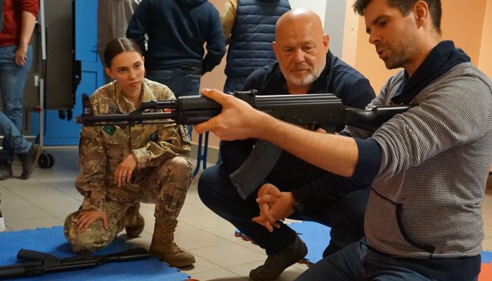 mężczyzna klęczy, trzyma karabin. obok kuca mężczyzna i dziewczyna w mundurze